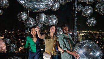 Group of three enjoying the reflective balloon room at SUMMIT One Vanderbilt with the NYC skyline in the background.