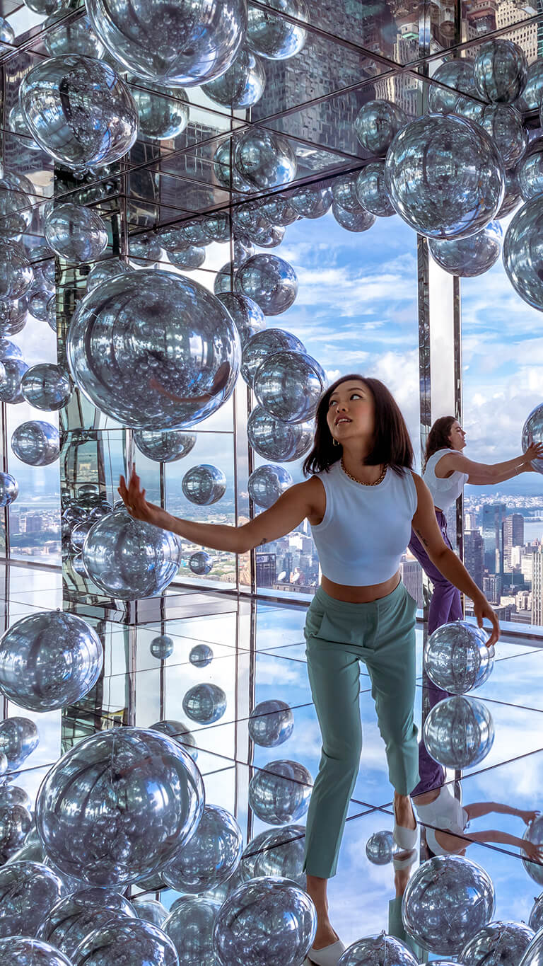Visitors in the mirror room with silver balloons, reflecting themselves against the New York City skyline backdrop.