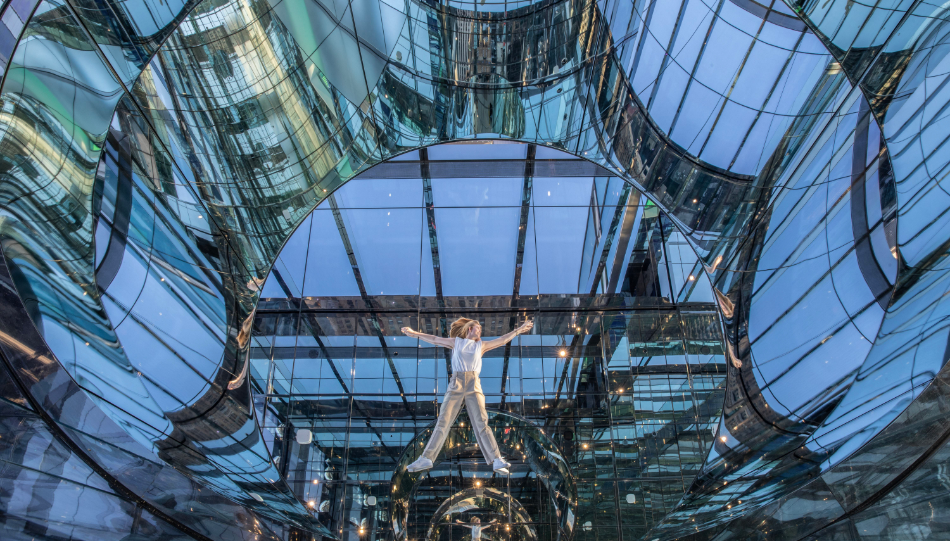 A person lying down in an interactive art exhibit in NYC, surrounded by curved, reflective surfaces.