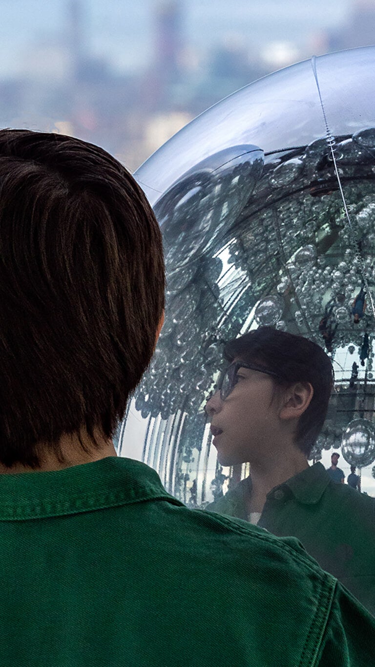 Young person in green shirt gazing into a large mirror ball, reflecting the New York City skyline in the background.