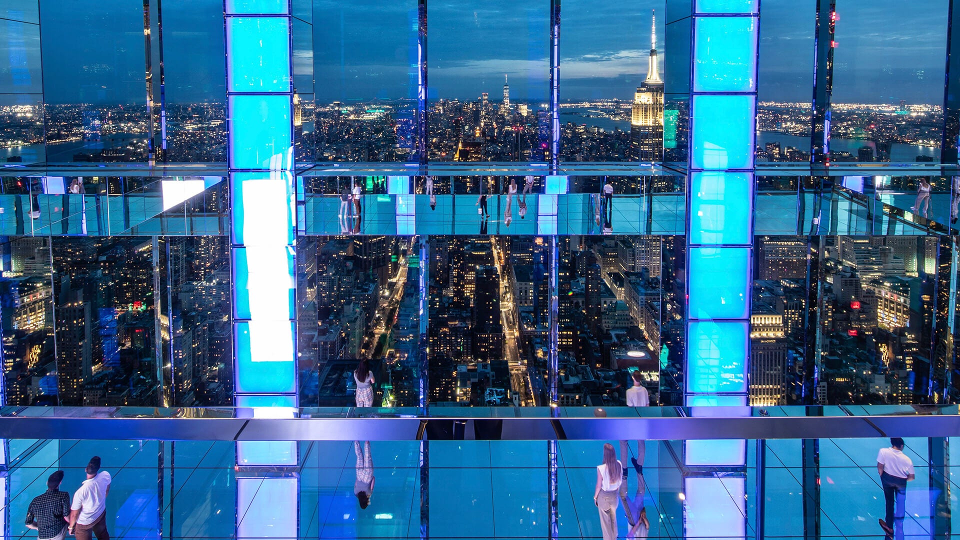 New York City night view from an observation deck with blue panels, showcasing the skyline and visitors.