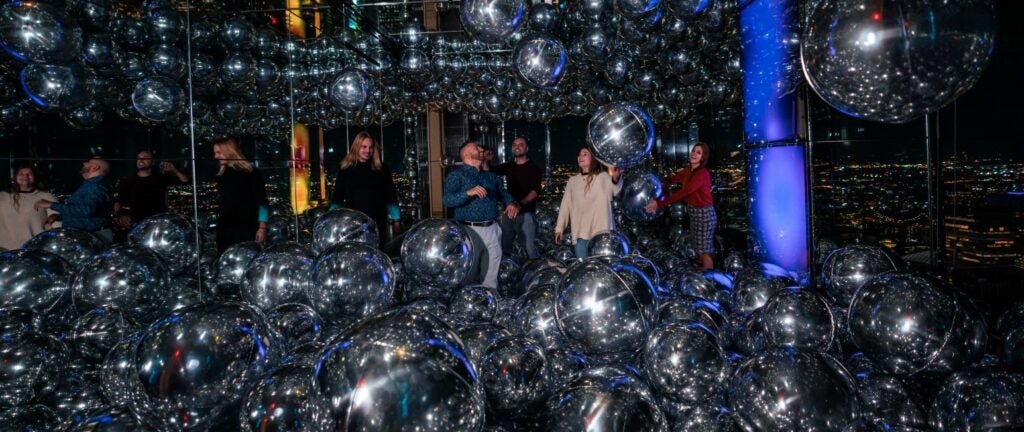 Visitors enjoy a fun corporate event surrounded by reflective silver balloons with a city night view.