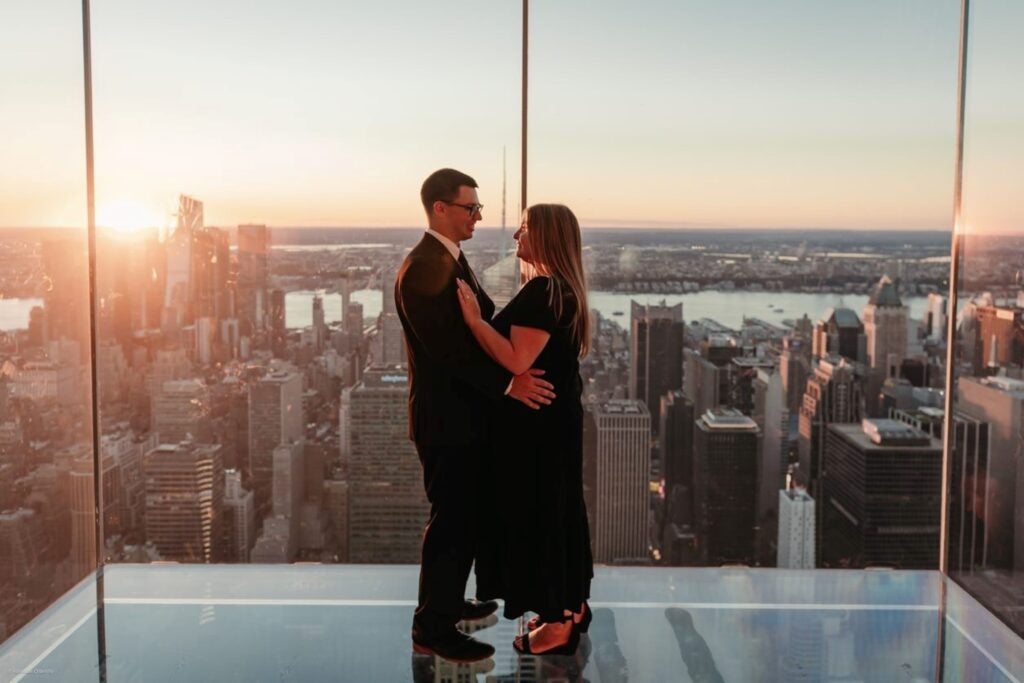 A couple embracing at a romantic proposal spot in NYC, with the city's sunset skyline in the background.