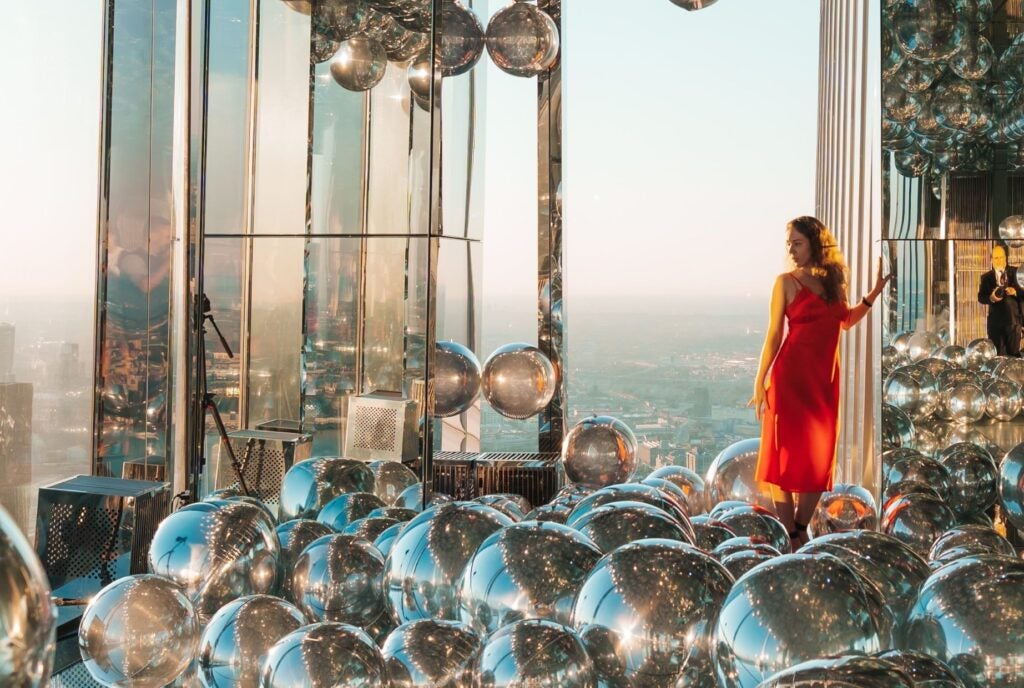 A woman in a red dress stands amidst reflective spheres at SUMMIT, offering a unique photo op with stunning views.
